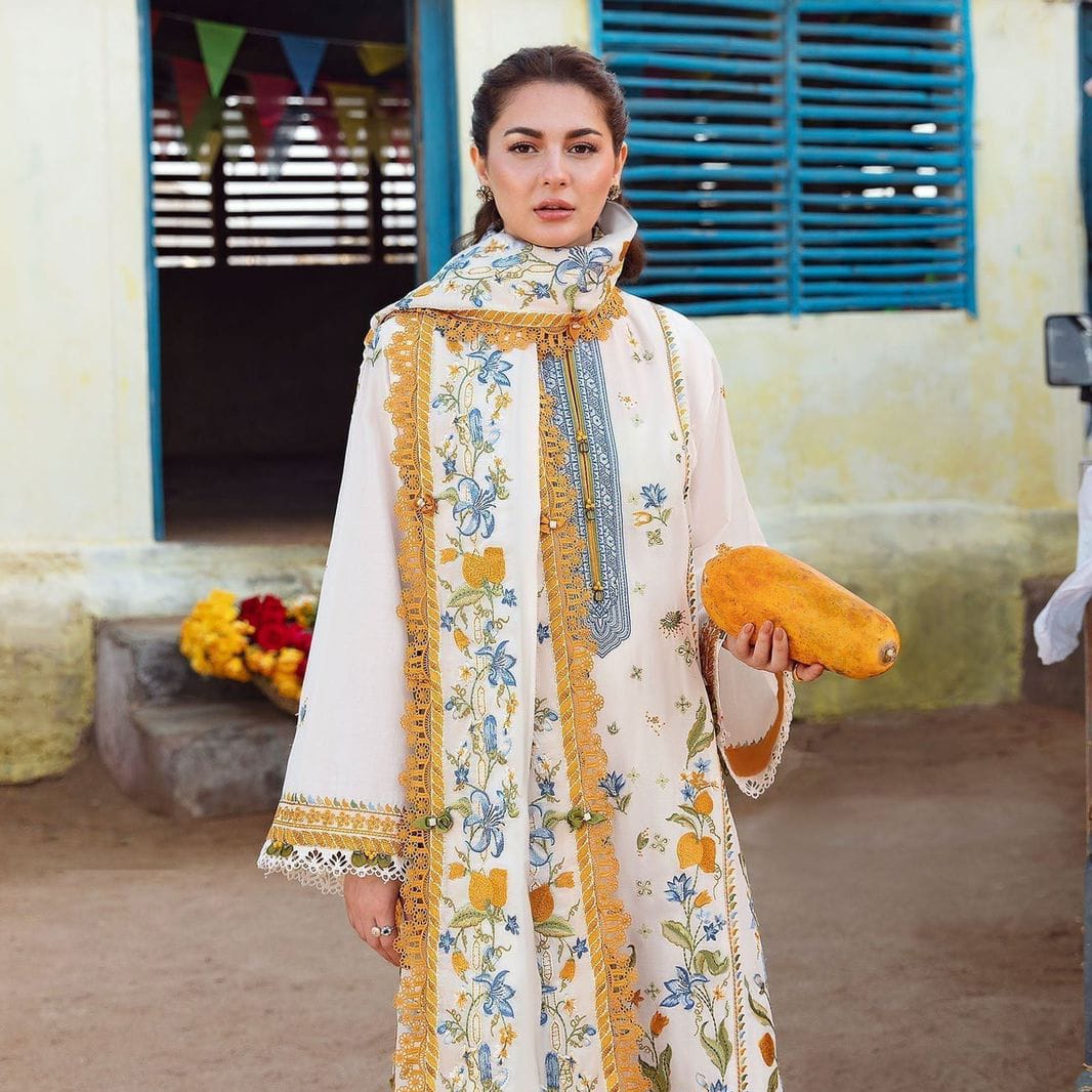 Model posing in an off-white embroidered luxury lawn 3 piece suit featuring mustard and blue floral patterns.