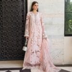 alt="A woman posing in a pastel pink luxury Pakistani formal dress with intricate floral embroidery, holding a silver embellished handbag and matching dupatta, standing against a tiled wall and decorative wooden door."