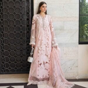 alt="A woman posing in a pastel pink luxury Pakistani formal dress with intricate floral embroidery, holding a silver embellished handbag and matching dupatta, standing against a tiled wall and decorative wooden door."