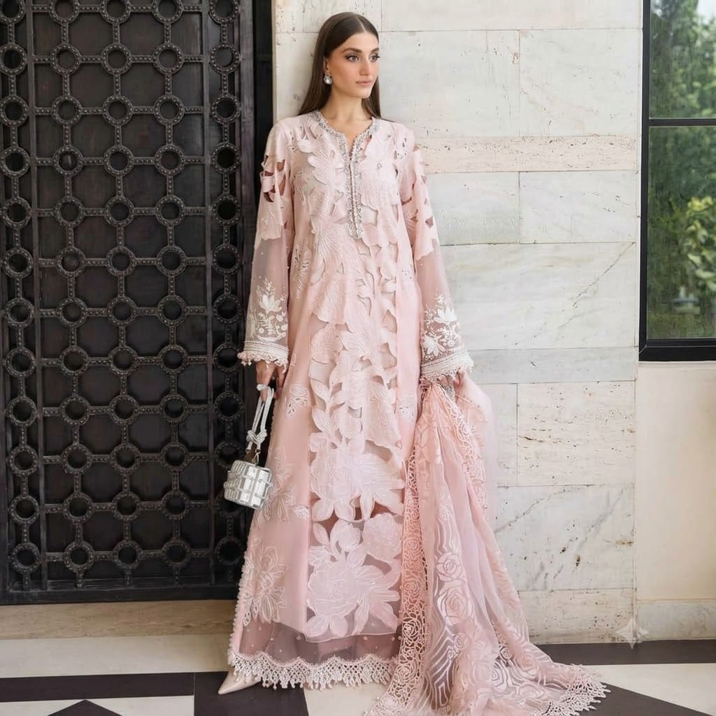 alt="A woman posing in a pastel pink luxury Pakistani formal dress with intricate floral embroidery, holding a silver embellished handbag and matching dupatta, standing against a tiled wall and decorative wooden door."